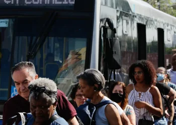Passageiros descendo de ônibus lotado em um ponto de transporte público movimentado durante o dia.
