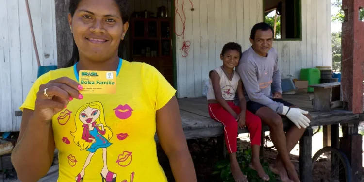 Mulher segurando um cartão do programa Bolsa Família em frente à sua casa, com um homem e uma criança ao fundo.