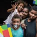 Crianças e uma mulher sorrindo em uma casa simples, com destaque para o cartão do Programa Bolsa Família.