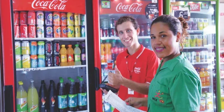 Dois funcionários sorrindo ao lado de refrigeradores Coca-Cola, verificando estoque de bebidas.