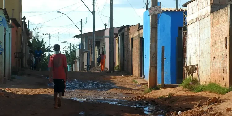 Rua de terra em uma comunidade com casas simples e moradores caminhando.