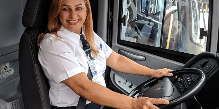 Motorista de ônibus sorridente ao volante, vestindo uniforme e cinto de segurança.