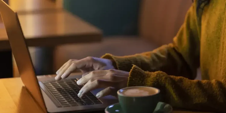 Mulher digitando no laptop enquanto toma café em ambiente aconchegante.