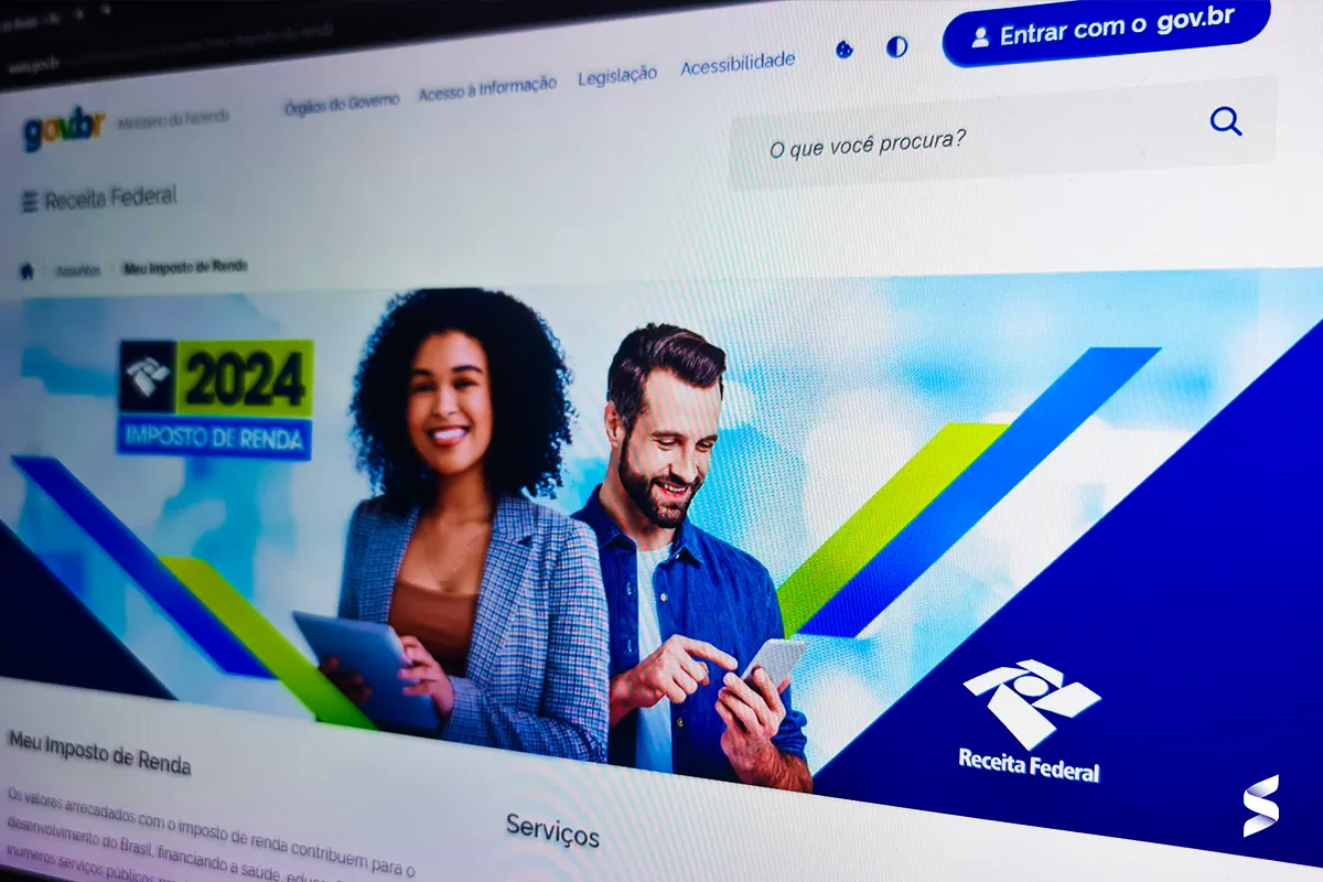 Página do site da Receita Federal com informações sobre o Imposto de Renda 2024, exibindo duas pessoas interagindo com dispositivos móveis.