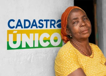 Mulher idosa com lenço laranja encostada em uma parede com o logo do Cadastro Único.