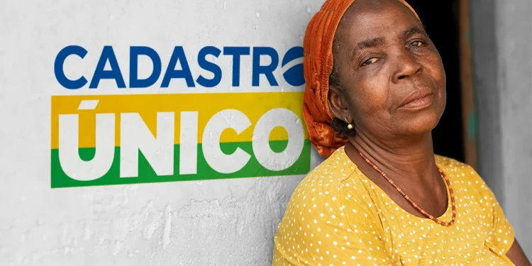 Mulher idosa com lenço laranja encostada em uma parede com o logo do Cadastro Único.