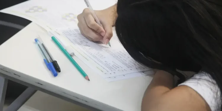 Pessoa fazendo uma prova, com a cabeça apoiada sobre a mesa e segurando um lápis enquanto escreve.