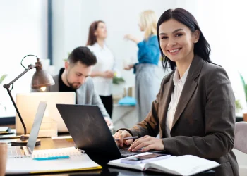 Mulher sorrindo enquanto trabalha em um laptop em um escritório moderno e colaborativo.