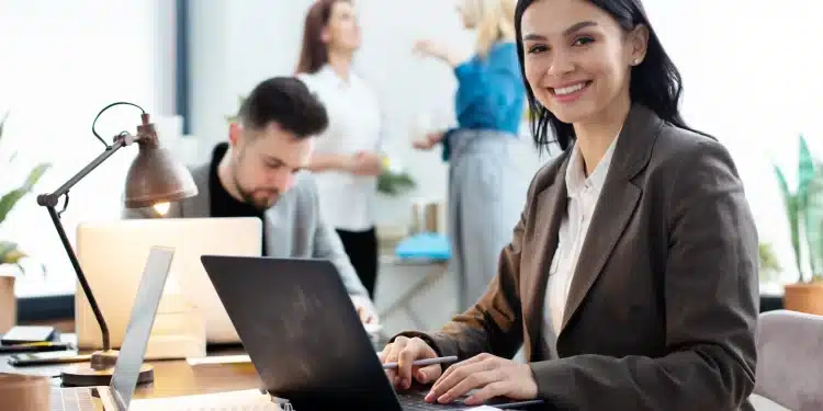 Mulher sorrindo enquanto trabalha em um laptop em um escritório moderno e colaborativo.