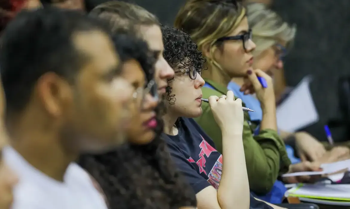 Grupo de pessoas atentas em uma palestra, com foco em estudante segurando caneta.