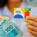 Cartão do Bolsa Família sendo segurado por uma pessoa, com cédulas de dinheiro ao fundo.