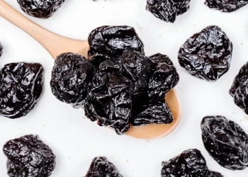 Ameixas secas em colher de madeira sobre fundo branco – alimento rico em fibras e antioxidantes.