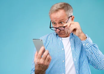 Homem mais velho ajustando os óculos enquanto observa um smartphone com expressão de curiosidade.