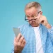 Homem mais velho ajustando os óculos enquanto observa um smartphone com expressão de curiosidade.