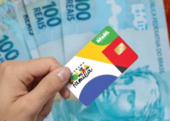 Cartão do programa social Bolsa Família 2025 sendo segurado por uma mão, com cédulas de dinheiro ao fundo.