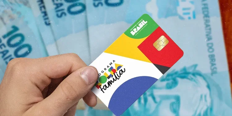Cartão do programa social Bolsa Família 2025 sendo segurado por uma mão, com cédulas de dinheiro ao fundo.