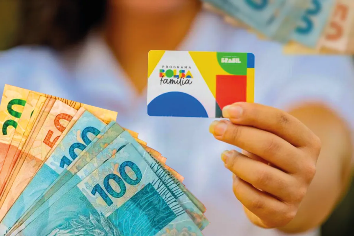 Cartão do Programa Bolsa Família segurado por uma mão, com cédulas de real ao fundo.