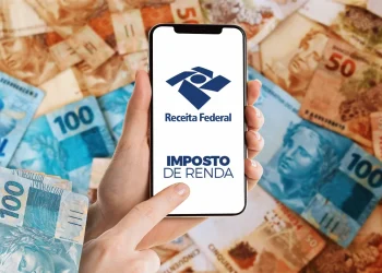 Celular exibindo a tela do site da Receita Federal com a inscrição "Imposto de Renda", sobre uma superfície coberta de notas de real.