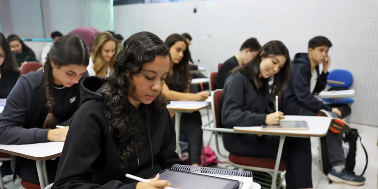 Estudantes concentrados em sala de aula, utilizando cadernos e tablets.