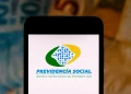 Logo da Previdência Social exibido na tela de um smartphone, com notas de dinheiro ao fundo, relacionado ao tema de verificação de descontos indevidos no INSS.