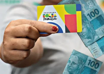 Pessoa segurando cartão do Bolsa Família com cédulas de 100 reais ao fundo