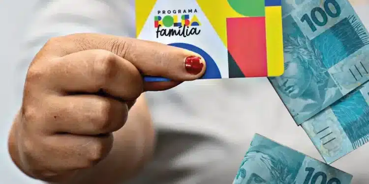 Pessoa segurando cartão do Bolsa Família com cédulas de 100 reais ao fundo