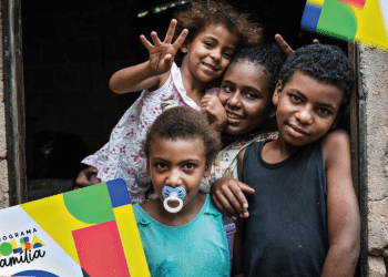 Quatro crianças sorrindo, em uma casa simples, com a bandeira do Programa Bolsa Família visível. Uma criança segura um cartão do programa, e outra usa um chupeta