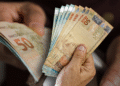Cédulas de reais sendo manuseadas por mãos idosas, simbolizando o recebimento de benefícios sociais