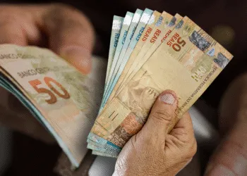 Cédulas de reais sendo manuseadas por mãos idosas, simbolizando o recebimento de benefícios sociais