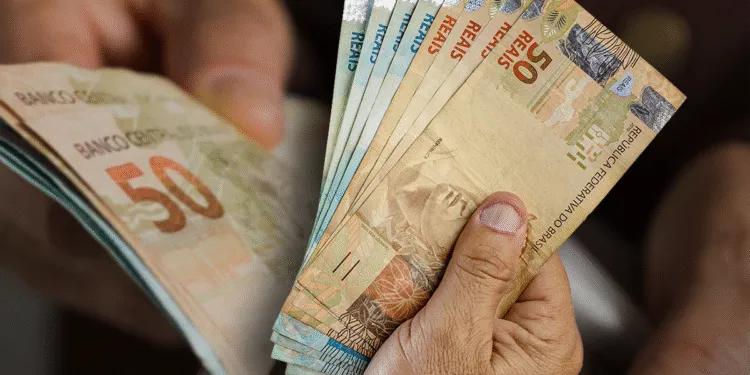 Cédulas de reais sendo manuseadas por mãos idosas, simbolizando o recebimento de benefícios sociais