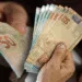 Cédulas de reais sendo manuseadas por mãos idosas, simbolizando o recebimento de benefícios sociais