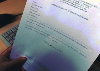Mão segurando um "Atestado de Antecedentes Criminais" com dados borrados, representando a solicitação legal deste certificado em processos de admissão e concursos públicos, como o da Polícia Federal.