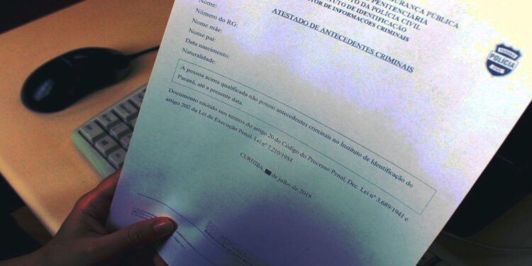 Mão segurando um "Atestado de Antecedentes Criminais" com dados borrados, representando a solicitação legal deste certificado em processos de admissão e concursos públicos, como o da Polícia Federal.