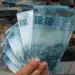 Mão segurando várias cédulas de 100 reais com grande quantidade de dinheiro amarrado em pacotes ao fundo