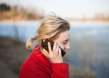 Jovem mulher loira, em área externa, falando ao celular, simbolizando a necessidade contínua de fazer ligações interurbanas e entender o código DDD, para ligar para outro estado.