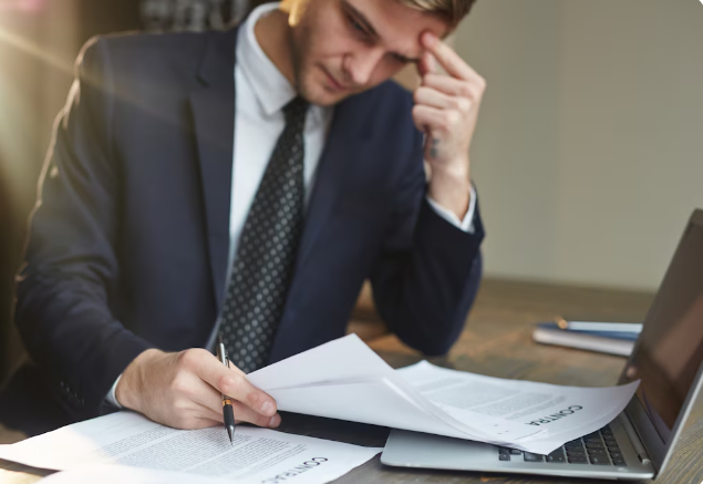 Homem vestido com terno azul analisando documentos com contrato próximo a computador em mesa de madeira