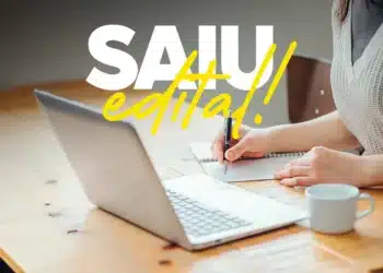 Pessoa anotando em caderno ao lado de notebook com texto 'SAIU edital!'