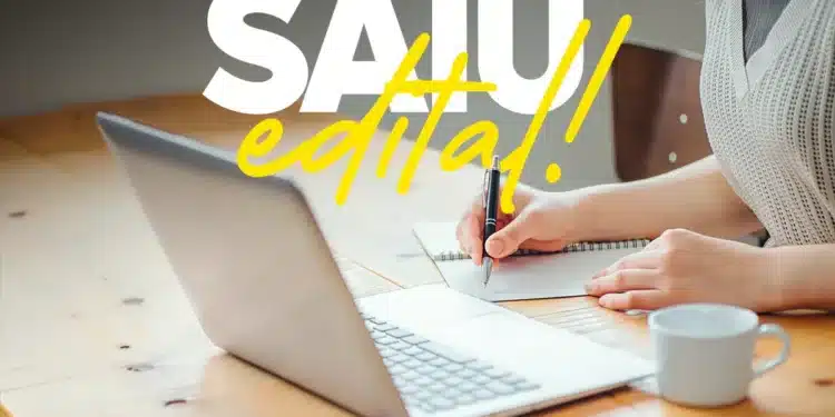 Pessoa anotando em caderno ao lado de notebook com texto 'SAIU edital!'