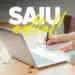 Pessoa anotando em caderno ao lado de notebook com texto 'SAIU edital!'