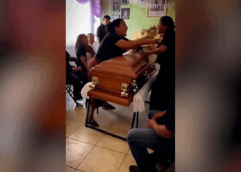 Duas mulheres discutem frente a frente sobre caixão marrom em velório , com pessoas ao fundo assistindo