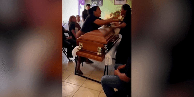 Duas mulheres discutem frente a frente sobre caixão marrom em velório , com pessoas ao fundo assistindo