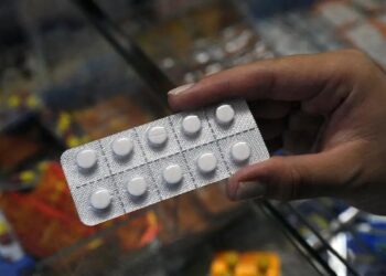 Mão enluvada segura ampola de dipirona monoidratada 500mg/mL com caixas do medicamento Hypofarma sobre a mesa.