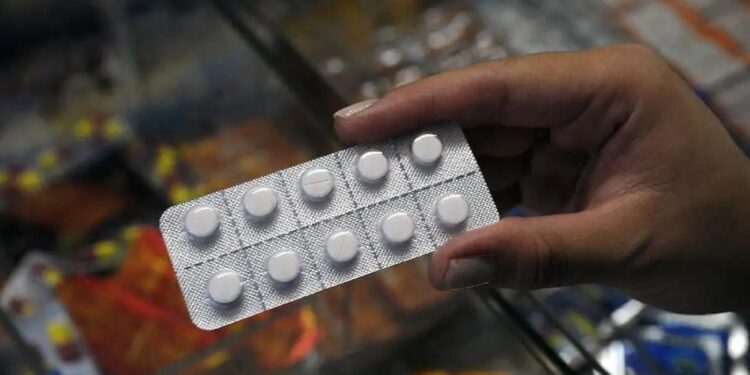 Mão enluvada segura ampola de dipirona monoidratada 500mg/mL com caixas do medicamento Hypofarma sobre a mesa.