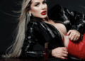 Mulher loira de batom vermelho com jaqueta preta e bolsa vermelha deitada ao lado de perfume
