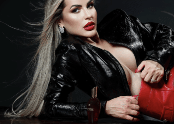 Mulher loira de batom vermelho com jaqueta preta e bolsa vermelha deitada ao lado de perfume