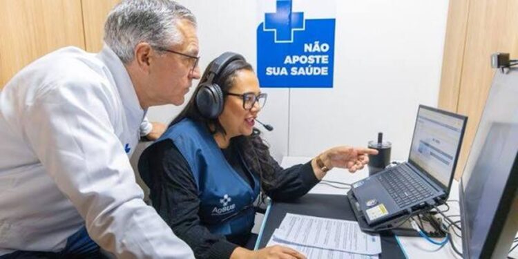 Atendente de headset e colete apontando para a tela de um notebook enquanto um homem acompanha ao lado, com o cartaz Não Aposte Sua Saúde ao fundo, referência ao apoio a pessoas afetadas por jogos e apostas